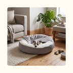 Petit chien endormi dans un panier apaisant gris posé sur un parquet, dans un salon lumineux